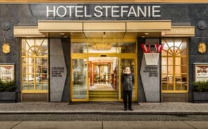 Lehrlingsportal.at Der Eingang des Hotels Stefanie, vor dem ein uniformierter Mitarbeiter steht, flankiert von Fenstern und goldverzierten Schildern.