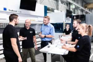 Lehrlingsportal.at Fünf Personen, vier in schwarzen Uniformen und eine in einem blauen Hemd, diskutieren an einem Tisch mit Papieren und Metallteilen in einem industriellen Arbeitsraum.