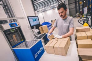 Lehrlingsportal.at Ein Lagerarbeiter mit Handschuhen und einem Arvato-Hemd verschließt Kartons an einer Packstation, in deren Nähe sich ein Computer und ein blauer Versandbehälter befinden.