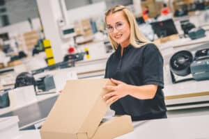 Lehrlingsportal.at Eine Frau mit schwarzem Hemd und Brille lächelt, während sie in einem modernen Arbeitsraum einen Karton zusammenbaut oder verpackt.