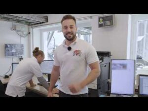 Lehrlingsportal.at Zwei Männer in einem modernen Büro mit Computerbildschirmen; einer steht und lächelt in die Kamera, der andere arbeitet an einem Schreibtisch.
