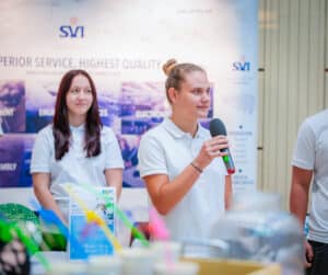 Lehrlingsportal.at Eine junge Frau in einem weißen Polohemd spricht in ein Mikrofon, während eine andere Frau in der Nähe steht, beide vor einem SVI-Werbebanner bei einer Indoor-Veranstaltung.