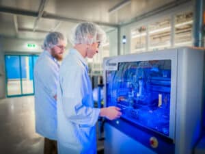 Lehrlingsportal.at Zwei Personen in Laborkitteln und Haarnetzen bedienen Maschinen in einem Labor mit blauer Beleuchtung.