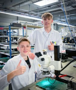 Lehrlingsportal.at Zwei junge Techniker in Laborkitteln zeigen neben einem Mikroskop und einer elektronischen Leiterplatte in einer hellen, organisierten Laborumgebung den Daumen nach oben.