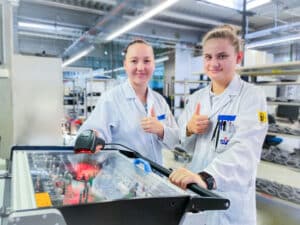 Lehrlingsportal.at Zwei Personen in Laborkitteln zeigen die Daumen nach oben, während sie in einer hellen, modernen Laborumgebung mit elektronischen Geräten arbeiten.