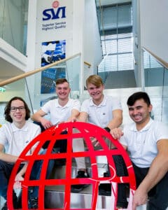 Lehrlingsportal.at Vier junge Männer in weißen Polohemden sitzen lächelnd auf einer Treppe im Innenbereich und halten eine große rote Weltkugel in der Hand. An der Wand hinter ihnen ist der Schriftzug der Firma SVI zu sehen.