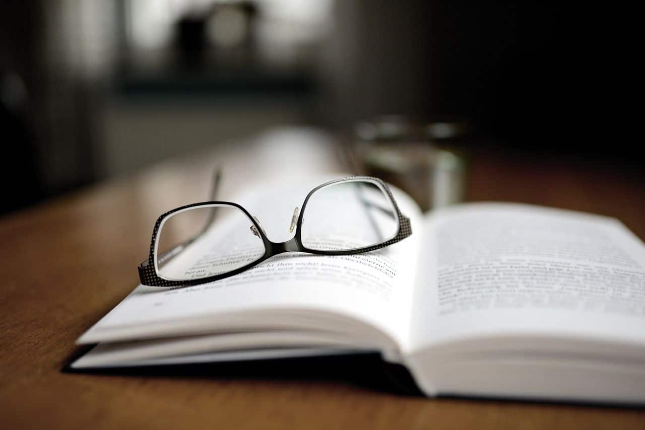 Lehrlingsportal.at Eine Brille ruht auf einem aufgeschlagenen Buch auf einem Holztisch, auf dem ein Glas Wasser und ein verschwommener Hintergrund zu sehen sind.
