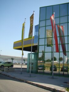 Lehrlingsportal.at Modernes Glasgebäude mit gelben Akzenten, mehrere Fahnen auf Masten und parkende Autos im Hintergrund an einem sonnigen Tag.