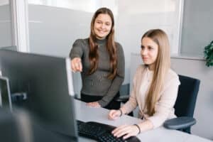 Lehrlingsportal.at Zwei Frauen in einem Büro blicken auf einen Computermonitor; die eine sitzt am Schreibtisch und benutzt den Computer, die andere steht daneben und zeigt auf den Bildschirm.