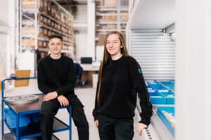 Lehrlingsportal.at Zwei junge Menschen in schwarzer Kleidung stehen in einem Lager oder einer Werkstatt, einer lehnt an einem Regal mit blauen Behältern, der andere sitzt auf einem Rollwagen.