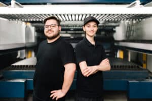 Lehrlingsportal.at Zwei Personen stehen nebeneinander in einer Fabrikhalle, beide tragen schwarze Hemden. Der eine hat eine Brille und einen Bart, der andere trägt eine Mütze und hat die Arme verschränkt. Im Hintergrund sind Industriemaschinen zu sehen.