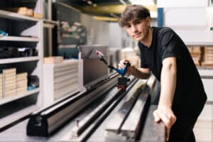Lehrlingsportal.at Ein junger Mann bedient in einer Werkstatt eine Werkzeugmaschine mit einem Handgerät und steht neben einer großen Metallschiene auf einer Werkbank.