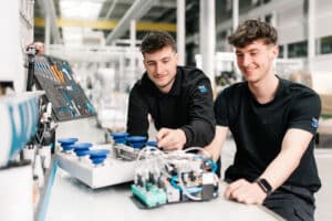 Lehrlingsportal.at Zwei junge Männer in schwarzen Hemden arbeiten mit elektronischen Bauteilen und Maschinen an einem Arbeitsplatz in einer modernen Industrieumgebung.