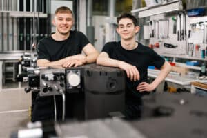 Lehrlingsportal.at Zwei junge Männer in schwarzen Hemden stehen in einer Werkstatt und stützen sich auf Industriegeräte, im Hintergrund sind Werkzeuge und Maschinen zu sehen.