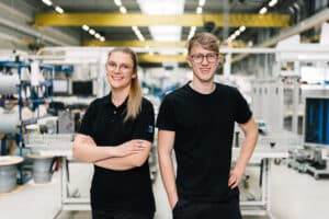 Lehrlingsportal.at Zwei junge Erwachsene mit Brillen und schwarzen Hemden stehen lächelnd in einer hellen Industrie- oder Produktionsanlage, umgeben von Maschinen und Geräten.
