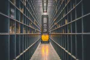 Lehrlingsportal.at Ein gelber Gabelstapler fährt durch einen schmalen Gang zwischen hohen Regalen in einem Lagerhaus, dessen Regale mit Kisten und Paketen gefüllt sind.