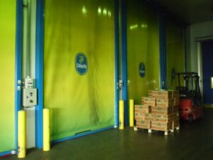 Lehrlingsportal.at Ein Gabelstapler und gestapelte Kisten stehen in einem Lagerhaus vor großen grünen und gelben Industrietoren mit Chiquita-Logos.