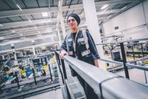 Lehrlingsportal.at Eine Person, die ein Kopftuch und eine Warnweste trägt, steht auf einer Plattform mit Blick auf eine industrielle Produktionsanlage.