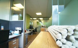 Lehrlingsportal.at Moderner Büroraum mit Schreibtischen, Stühlen und Computern, die durch Glastrennwände voneinander getrennt sind; ein aufgerollter weißer Teppich liegt auf einer Holzfläche im Vordergrund.