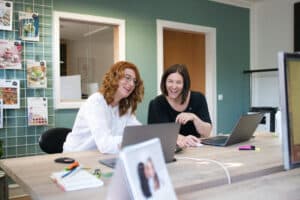 Lehrlingsportal.at Zwei Frauen sitzen an einem Schreibtisch in einem Büro, beide lächeln und schauen auf einen Laptop-Bildschirm; auf dem Tisch liegen Büromaterial und Papiere.