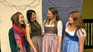 Lehrlingsportal.at Vier Frauen stehen zusammen, drei in traditionellen Dirndlkleidern und eine mit einem bunten Schal, lächeln und interagieren vor einer strukturierten Indoor-Kulisse mit Lichterketten.