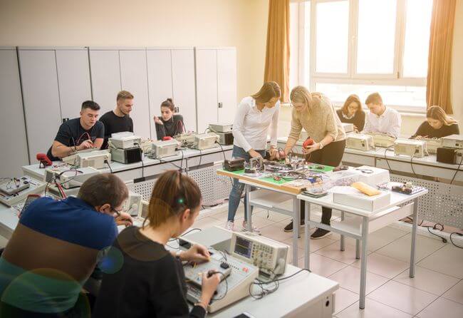 Lehrlingsportal.at In einem Klassenzimmer mit großen Fenstern und technischen Geräten arbeitet eine Gruppe von Schülerinnen und Schülern an Tischen mit elektronischen Geräten und Platinen.