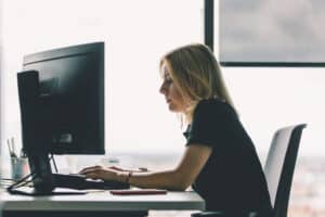 Lehrlingsportal.at Eine Frau sitzt an einem Schreibtisch und arbeitet an einem Computer in einem modernen Büro, durch ein großes Fenster hinter ihr fällt natürliches Licht.