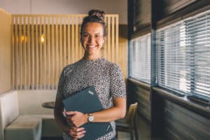 Lehrlingsportal.at Eine Frau, die eine Brille und ein gemustertes Oberteil trägt, steht in einem Innenraum, lächelt und hält einen geschlossenen Lenovo ThinkPad-Laptop in der Hand, während im Hintergrund Bürositze und große Fenster zu sehen sind.