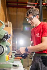 Lehrlingsportal.at Ein junger Mensch mit Schutzbrille und rotem Hemd arbeitet in einer Werkstatt an einer Schleifmaschine, mit hellem Licht im Hintergrund.