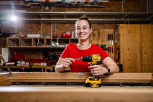 Lehrlingsportal.at Eine Person mit Brille und rotem Hemd hält lächelnd eine gelbe Bohrmaschine in der Hand und steht hinter einem Holzrahmen in einer Werkstatt.