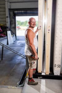 Lehrlingsportal.at Ein Mann in einem Tank-Top und Shorts steht in einem Innenraum neben Holzplatten, hält ein Elektrowerkzeug in der Hand und lächelt in die Kamera.