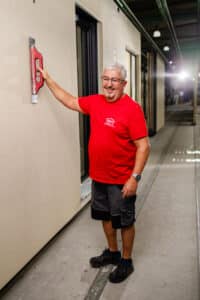 Lehrlingsportal.at Ein älterer Mann in einem roten T-Shirt und schwarzen Shorts glättet mit einer Kelle den Putz an einer Wand in einer Baustelle.