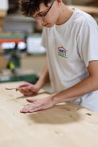 Lehrlingsportal.at Eine Person, die eine Brille und ein weißes T-Shirt trägt, arbeitet mit einem Werkzeug auf einer Holzoberfläche in einer Holzwerkstatt.