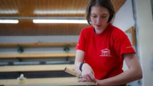 Lehrlingsportal.at Eine Person in einem roten Hartl-Haus-Hemd bearbeitet mit einem Handwerkzeug ein Stück Holz in einer Werkstatt.