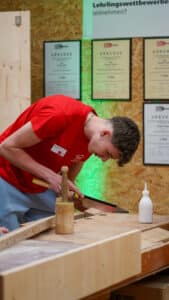 Lehrlingsportal.at Eine Person in einem roten Hemd sägt mit einer Handsäge an einem Stück Holz an einer Werkbank, in der Nähe befinden sich Tischlerwerkzeuge und eine Flasche Leim. An der Wand hinter ihnen hängen Urkunden aus.