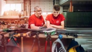 Lehrlingsportal.at Zwei Personen in roten Hemden stehen an einer Werkbank in einer Werkstatt, umgeben von Werkzeugen, unterhalten sich und schauen auf die Arbeitsfläche hinunter.