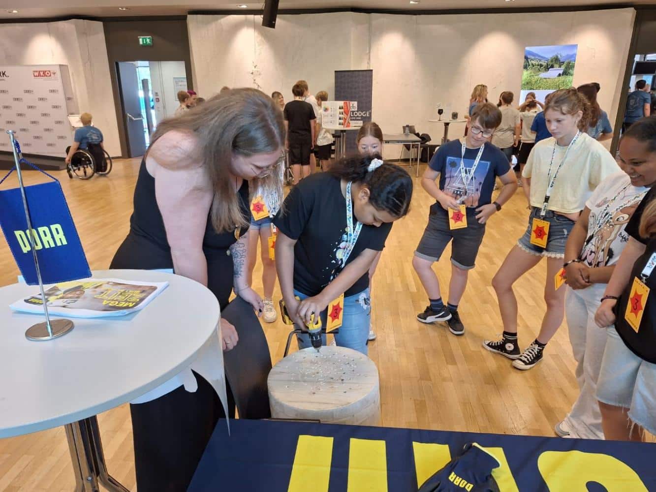 Lehrlingsportal.at Eine Gruppe von Kindern sieht zu, wie ein Mädchen unter Aufsicht eines Erwachsenen bei einer Indoor-Veranstaltung mit Informationsständen in einen Holzblock bohrt.