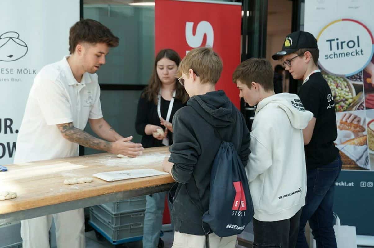 Lehrlingsportal.at Ein Mann führt einer Gruppe von drei Jungen und einem Mädchen an einem Tisch das Ausrollen von Teig vor, während im Hintergrund Werbebanner zu sehen sind.