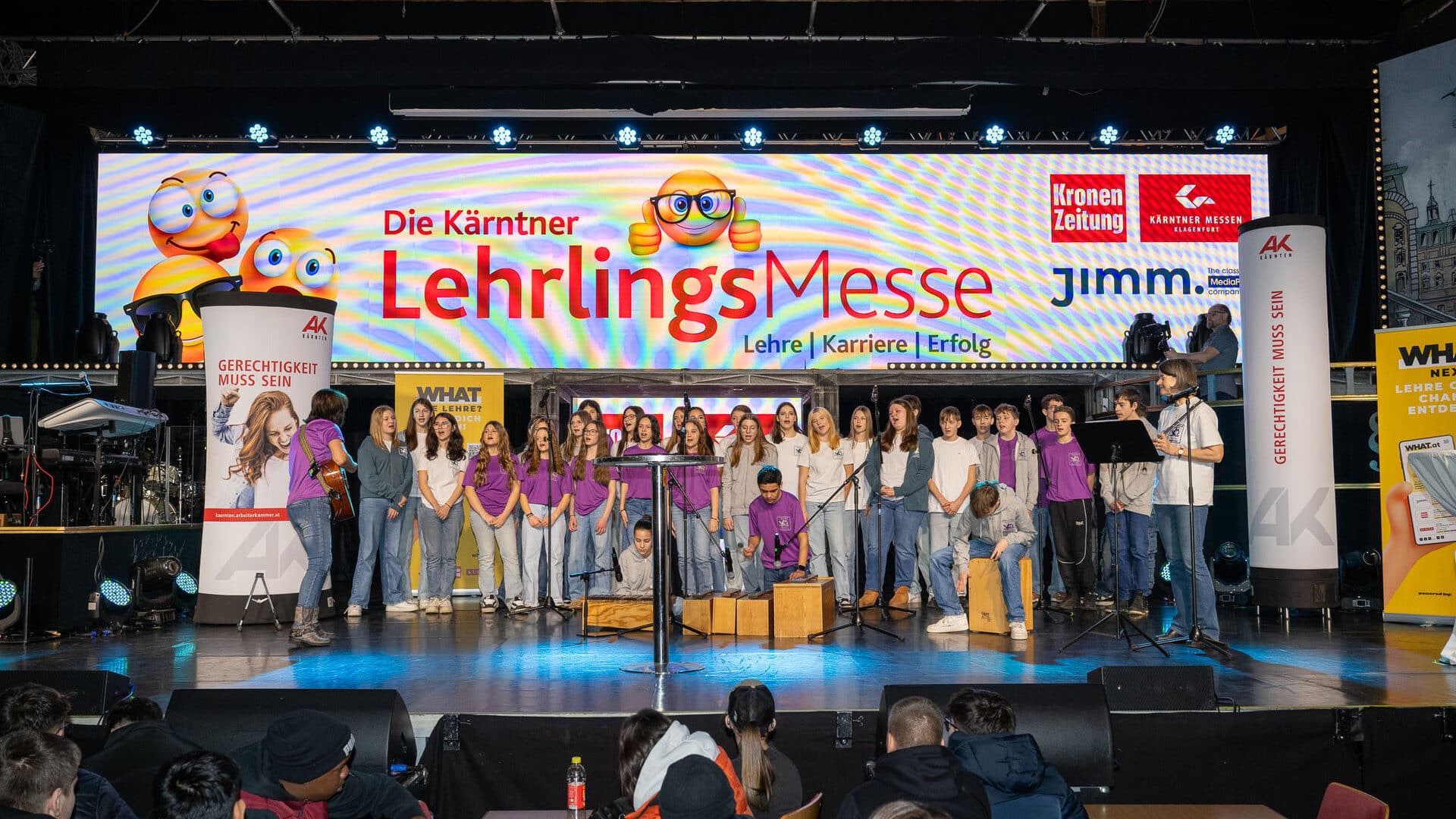 Lehrlingsportal.at Eine große Gruppe von Schülerinnen und Schülern musiziert auf der Bühne der Kärntner Lehrlingsmesse, davor sitzt ein buntes Transparent und das Publikum.