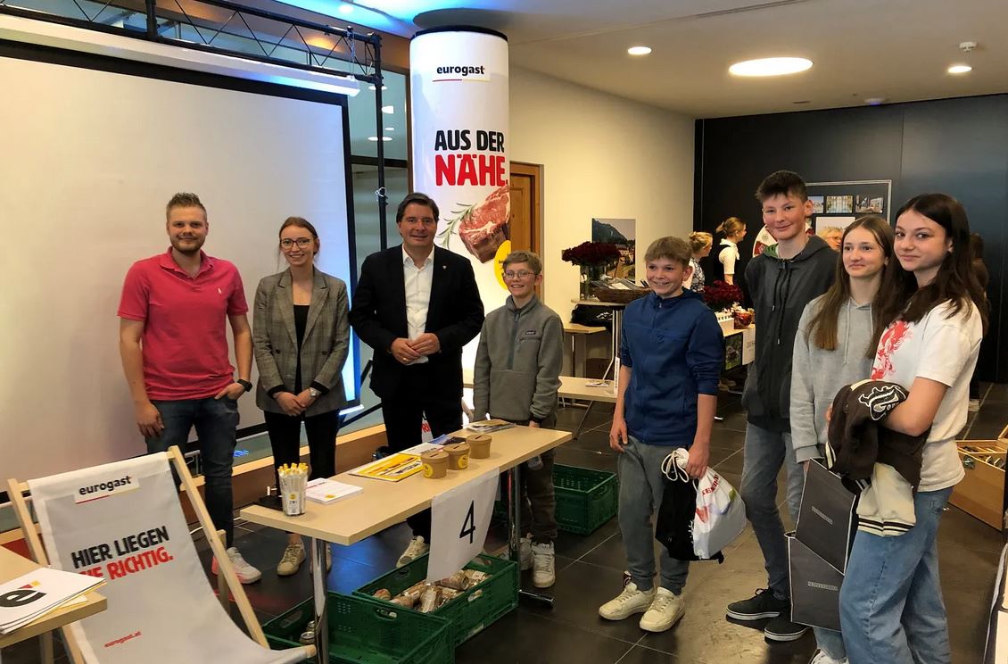 Lehrlingsportal.at Eine Gruppe von Erwachsenen und Jugendlichen steht lächelnd in der Nähe eines Ausstellungstisches bei einer Indoor-Veranstaltung, während im Hintergrund Werbeplakate und -materialien zu sehen sind.