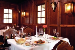 Lehrlingsportal.at Eleganter Restauranttisch für vier Personen mit weißem Tischtuch, Weingläsern, Tellern mit Speisen, einem Brotkorb, einer brennenden Kerze und holzgetäfelten Wänden mit montiertem Hirschgeweih und Laternen.