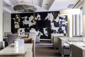 Lehrlingsportal.at Moderner Loungebereich mit gemusterten Sesseln, hellen Holztischen, großen Fenstern und einem Fernseher an einer schwarz-weißen Wand mit Kuhdruck.