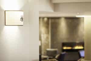 Lehrlingsportal.at Wandleuchte an einer weißen Wand im Vordergrund, mit einer unscharfen Sitzecke und einem Kamin im Hintergrund.