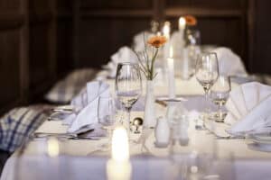 Lehrlingsportal.at Ein formeller Esstisch mit weißer Bettwäsche, Glaswaren, gefalteten Servietten, einer Vase mit einer orangefarbenen Blume und brennenden Kerzen in einem sanft beleuchteten Raum.
