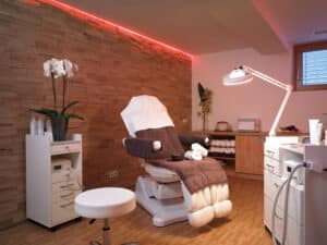 Lehrlingsportal.at Ein moderner Spa-Behandlungsraum mit einem Liegestuhl, weißen Möbeln, Handtüchern, einer Orchideenpflanze und sanfter Beleuchtung.