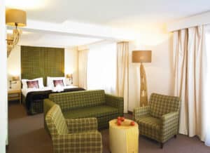 Lehrlingsportal.at Hotelzimmer mit einem Doppelbett, grün kariertem Sofa und Sesseln, einem kleinen Holztisch mit Äpfeln, neutralen Wänden und großen Fenstern mit beigen Vorhängen.