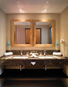 Lehrlingsportal.at Doppelwaschbecken mit Granitarbeitsplatte, zwei Spiegel in Holzrahmen, aufgerollte Handtücher und montierter Haartrockner unter Deckeneinbauleuchten.