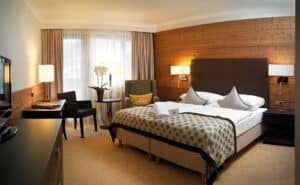 Lehrlingsportal.at Ein Hotelzimmer mit einem Doppelbett, einer Akzentwand aus Holz, einem Sessel, einem Schreibtisch mit Stuhl, zwei Nachttischlampen, einem Fenster mit Vorhängen und einer Topforchidee auf einem runden Tisch.