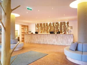 Lehrlingsportal.at Moderner Hotelempfangsbereich mit einem hölzernen Schreibtisch, zwei Mitarbeitern, dekorativer Holzvertäfelung, runden Sitzgelegenheiten und heller Beleuchtung.