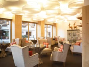 Lehrlingsportal.at Eine gemütliche Lounge mit cremefarbenen Sesseln, Glastischen, karierten Kissen, großen Fenstern, kunstvollen Beleuchtungskörpern und einem bemalten Schrank mit Hirschfiguren auf dem Kopf.